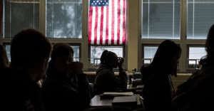 Students sit in a dimly lit classroom with an American flag hanging in front of a window, a subtle reminder of America's education problem.