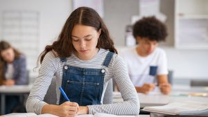 A student in a striped shirt and denim overalls writes diligently in a notebook, likely preparing for the 2025 SAT, with another student blurred in the background.