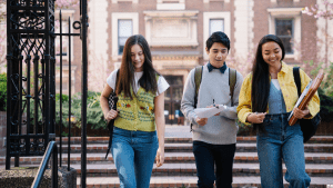 Three students with backpacks and books walk together on a university campus, with a brick building and blooming trees in the background.