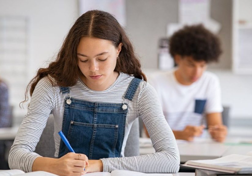 A student in a striped shirt and denim overalls writes diligently in a notebook, likely preparing for the 2025 SAT, with another student blurred in the background.