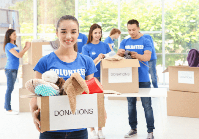 In a bright room, smiling volunteers in blue shirts, perhaps participating in extracurricular activities in high school, organize donation boxes filled with clothing and other items.