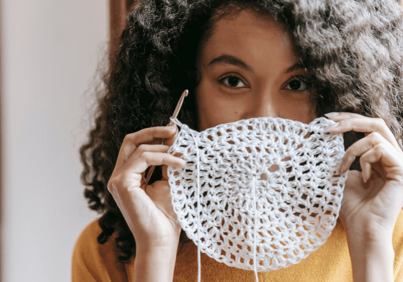 With curly hair cascading around their face, the person holds up a partially completed crochet piece, cleverly using their hobby not just for relaxation but as a unique element in crafting standout college applications.