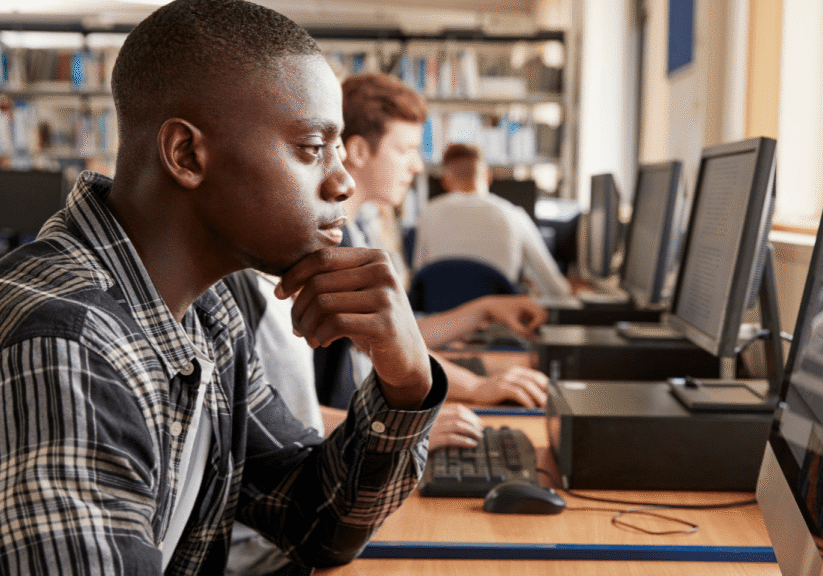 Several students sit at desks using desktop computers in a library or computer lab, focusing intently on their screens—perhaps researching how to build a college list. Bookshelves are visible in the background.