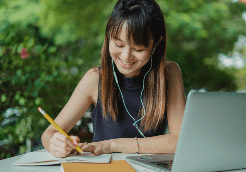 A person sits at an outdoor table, smiling while writing in a notebook with a pencil, wearing earphones, with a laptop open in front of them and greenery in the background.