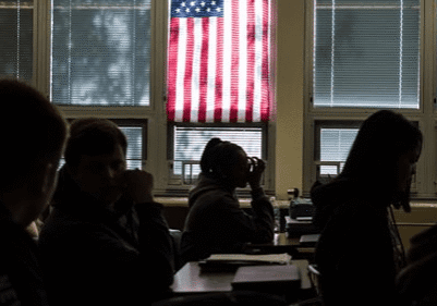 Students sit in a dimly lit classroom with an American flag hanging in front of a window, a subtle reminder of America's education problem.