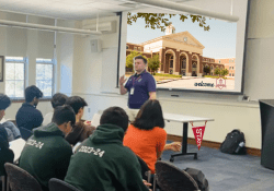 A person stands at the front of a classroom presenting to a seated audience, with a projection of a building on the screen behind them.