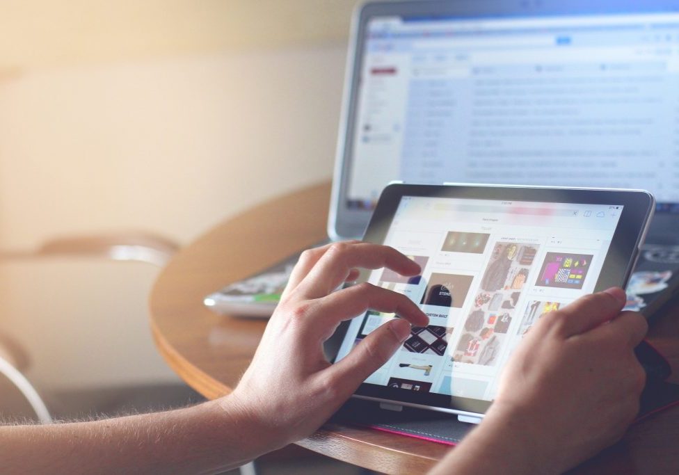 A person is using a tablet on a table, browsing images. A laptop in the background displays an open webpage with text and images. The scene is indoors with natural light streaming from the side, creating a serene atmosphere that suggests you might have missed it if not paying attention.